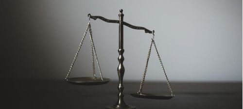 A bronze Scale of Justice stands in the center of the frame against a neutral, two-toned grey background. The scale is slightly unbalanced, with the left pan hanging marginally lower than the right.