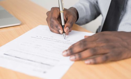 A close-up of a person wearing a grey dress shirt and black tie, sitting at a wooden desk and signing a document with a silver pen.