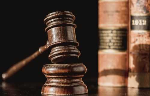  A wooden gavel and sound block sit on a dark surface in the foreground, with old, leather-bound law books standing in the blurred background against a dark setting.
